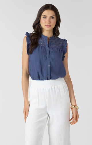 Woman wearing a blue sleeveless blouse with lace details and white pants on a white background