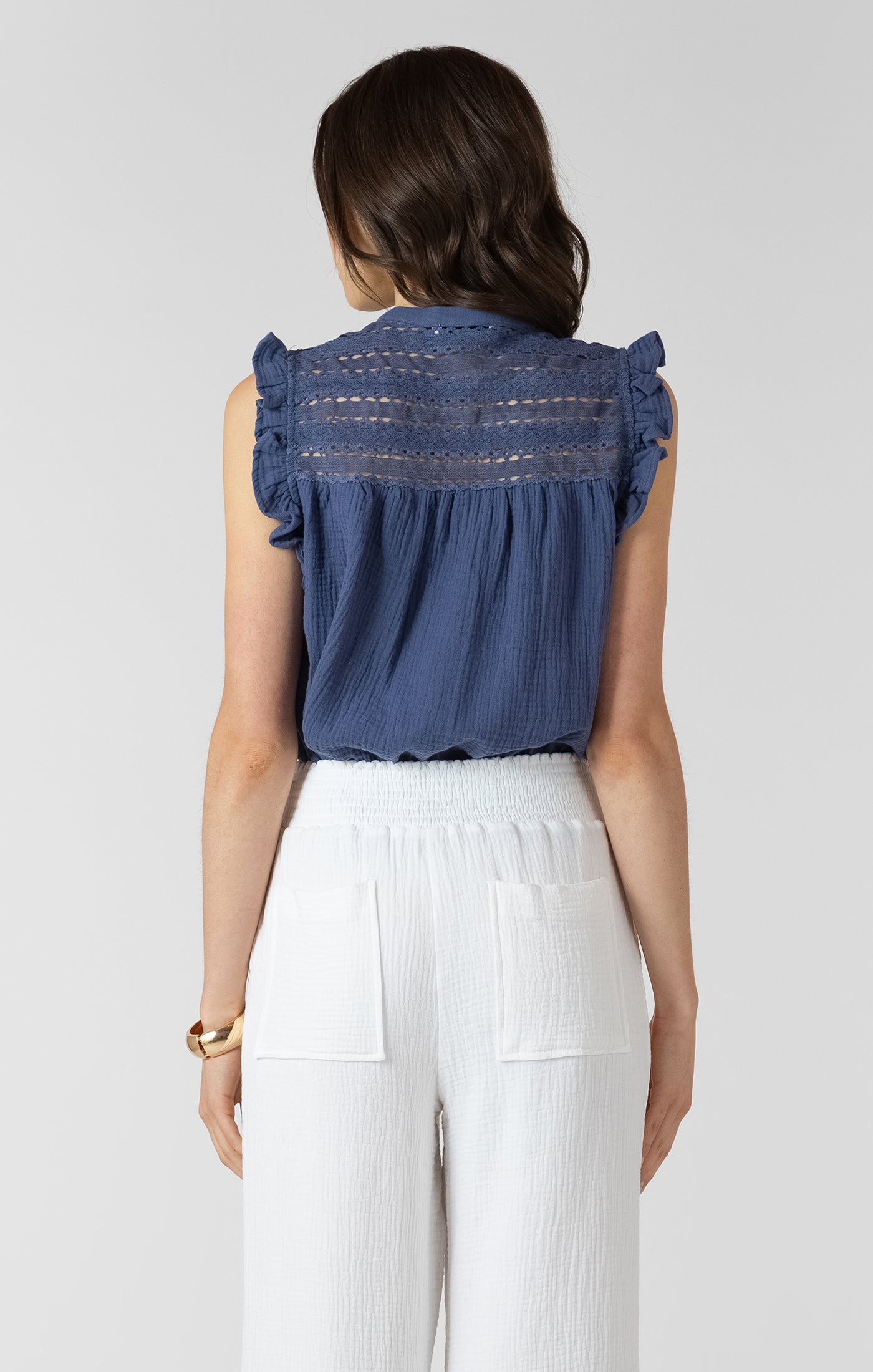 Woman wearing a blue sleeveless top with lace details and white pants on a plain background
