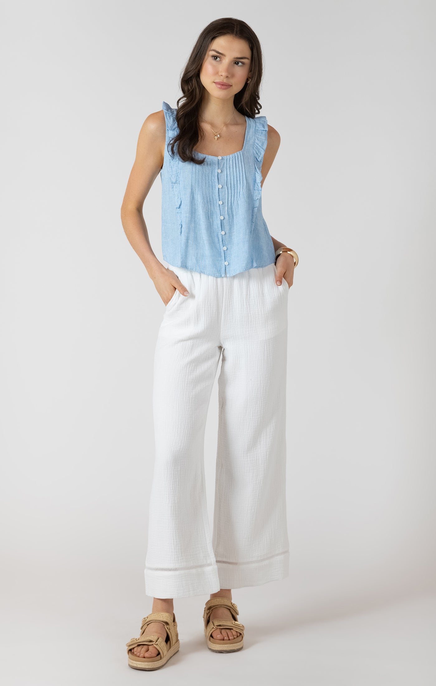 Woman wearing a light blue sleeveless top and white pants on a plain background