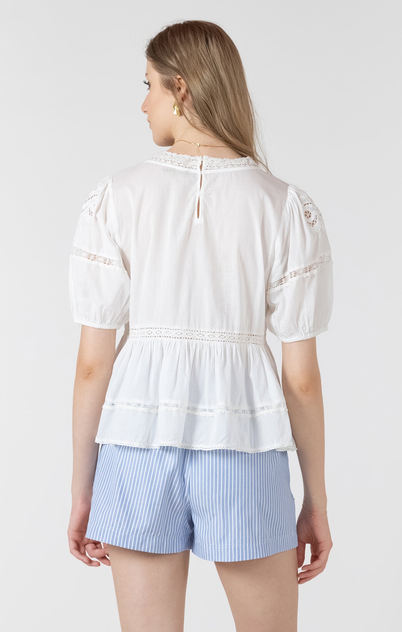 Woman wearing a white blouse with lace details and light blue striped shorts on a plain background