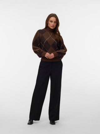 A woman modeling a brown argyle knit turtleneck sweater with black wide-leg trousers, standing against a light gray studio background.