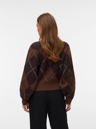 A woman viewed from behind wearing a brown argyle sweater with black pants, set against a plain white studio background.