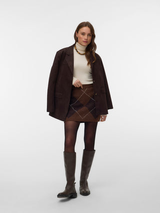 A model stands against a light gray studio background wearing a dark brown tailored blazer over a cream mock-neck top, a brown argyle mini skirt, dark tights, and dark brown knee-high boots.