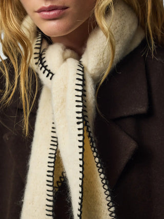 Close-up of a model wearing a thick cream-colored knit scarf with black zigzag contrast stitching, styled with a dark brown coat.