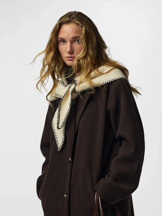 A woman wearing a dark brown wool coat with a cream-colored shearling-style collar and a matching tie, photographed against a light gray studio background.