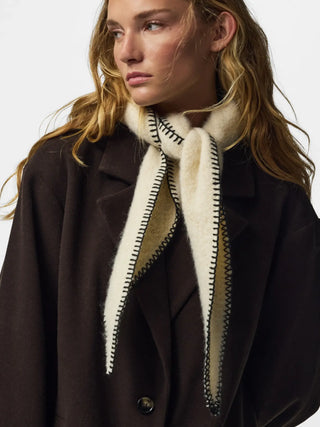 A close-up of a model wearing a dark brown coat and a cream-colored scarf with black zigzag stitching and a beige lining.