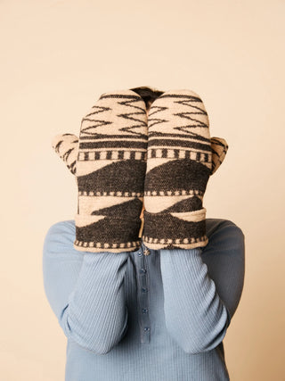 A person wearing a light blue ribbed long-sleeve top holds a pair of knit winter mittens in front of their face. The mittens have a Southwestern geometric pattern in beige and dark brown.