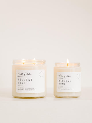 Two candles with labels reading 'Welcome Home', one larger with a single wick and one smaller with a double wick, set against a neutral background.