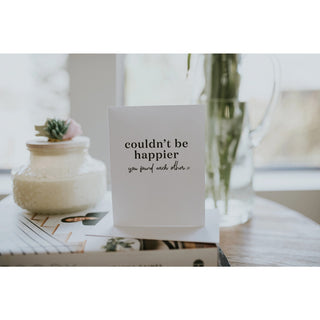Card with 'couldn't be happier you found each other' text on a table with a candle and plant.