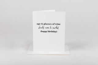 A greeting card with a humorous birthday message that reads 'Age & Wine should never be counted', printed in black ink. The card has a white background, a blank interior for a personal message, and a white Euro flap envelope.