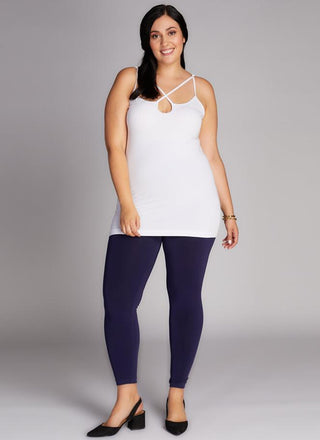 A woman wearing a white tank top and navy high-waisted leggings, standing against a grey background.