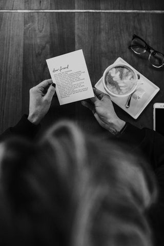 A woman holding a greeting card on a table with a coffee and reading glasses. 
