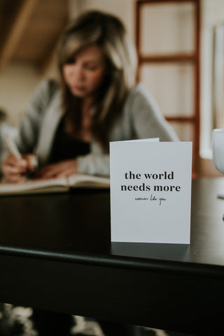 A greeting card with the text 'the world needs more women like you' placed on a table with a person writing in the background.