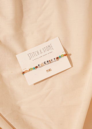 A colorful beaded bracelet with the phrase 'F*ck It' on a cardboard display, set against a beige background.