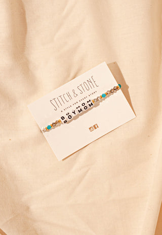 Bracelet with 'BOY MOM' on a card with 'Stitch & Stone' branding, on a beige fabric background