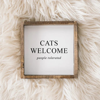 Wooden-framed sign with 'Cats Welcome, people tolerated' text on a fluffy white surface