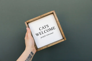 a hand holding a wooden sign that says "cats welcome, people tolerated"